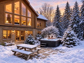 Une cabane en rondins avec un jacuzzi et une table de pique-nique en bois, le tout situé dans un paysage enneigé.