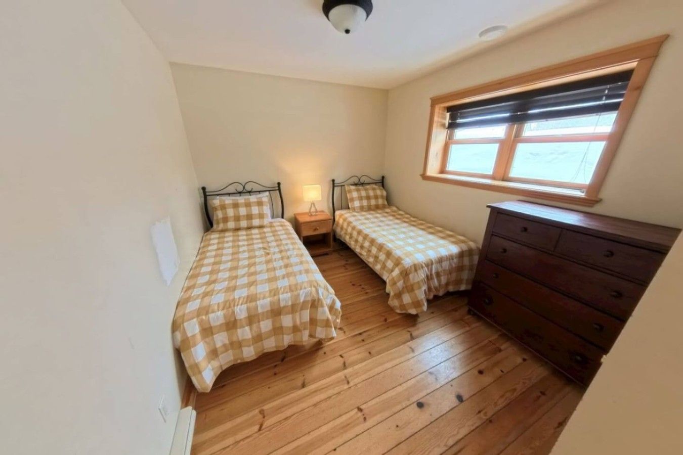 A small room with two single beds, a wooden dresser, a nightstand with a lamp, and a window with blinds.