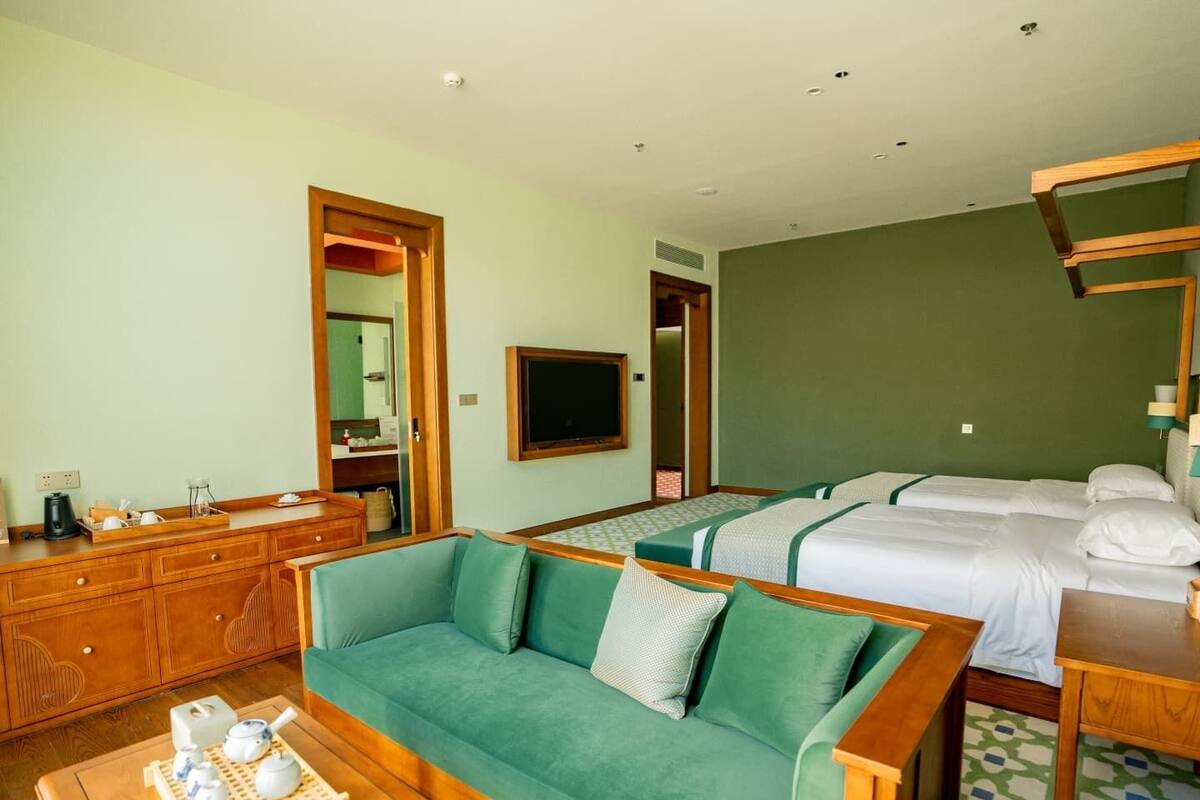 Habitación de hotel con sofá verde, cómoda de madera, cama con ropa blanca y televisor en la pared.