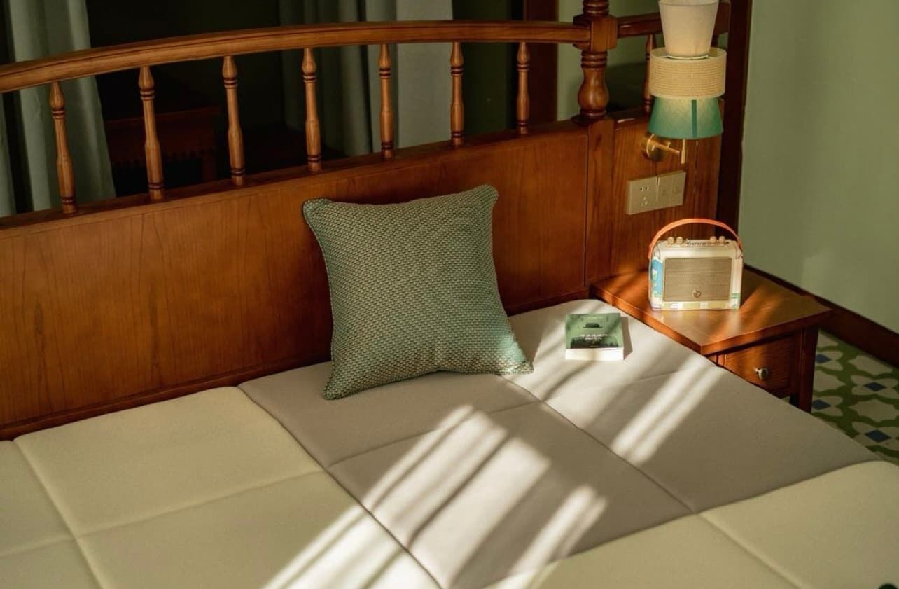 Una cama con cabecera de madera, un almohadón, una mesita de noche con un radio y una pared verde.