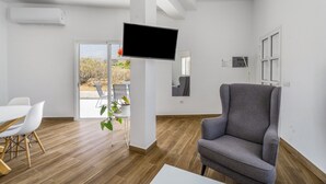 A modern room with a flat-screen TV, a grey armchair, a dining table with chairs, and a large window with a view of greenery.