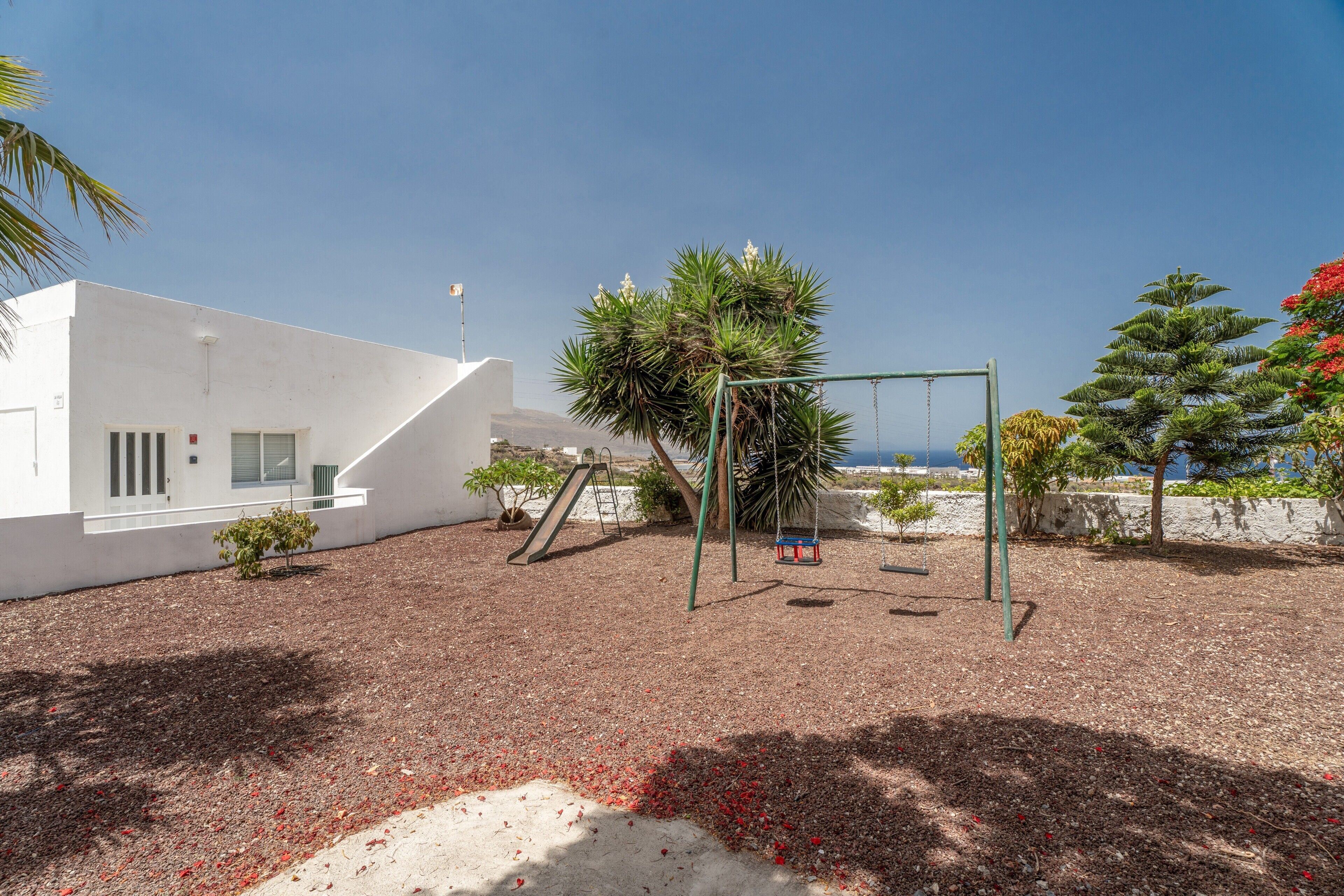 A playground with swings and a slide, surrounded by trees and a building with a white facade.