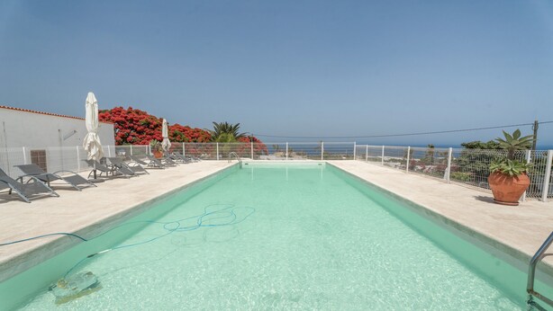 A rectangular swimming pool with clear water, surrounded by a white fence, and a view of the sea in the background.