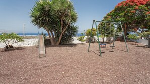 Un parque infantil con columpios y un tobogán, rodeado de árboles y vista al mar.