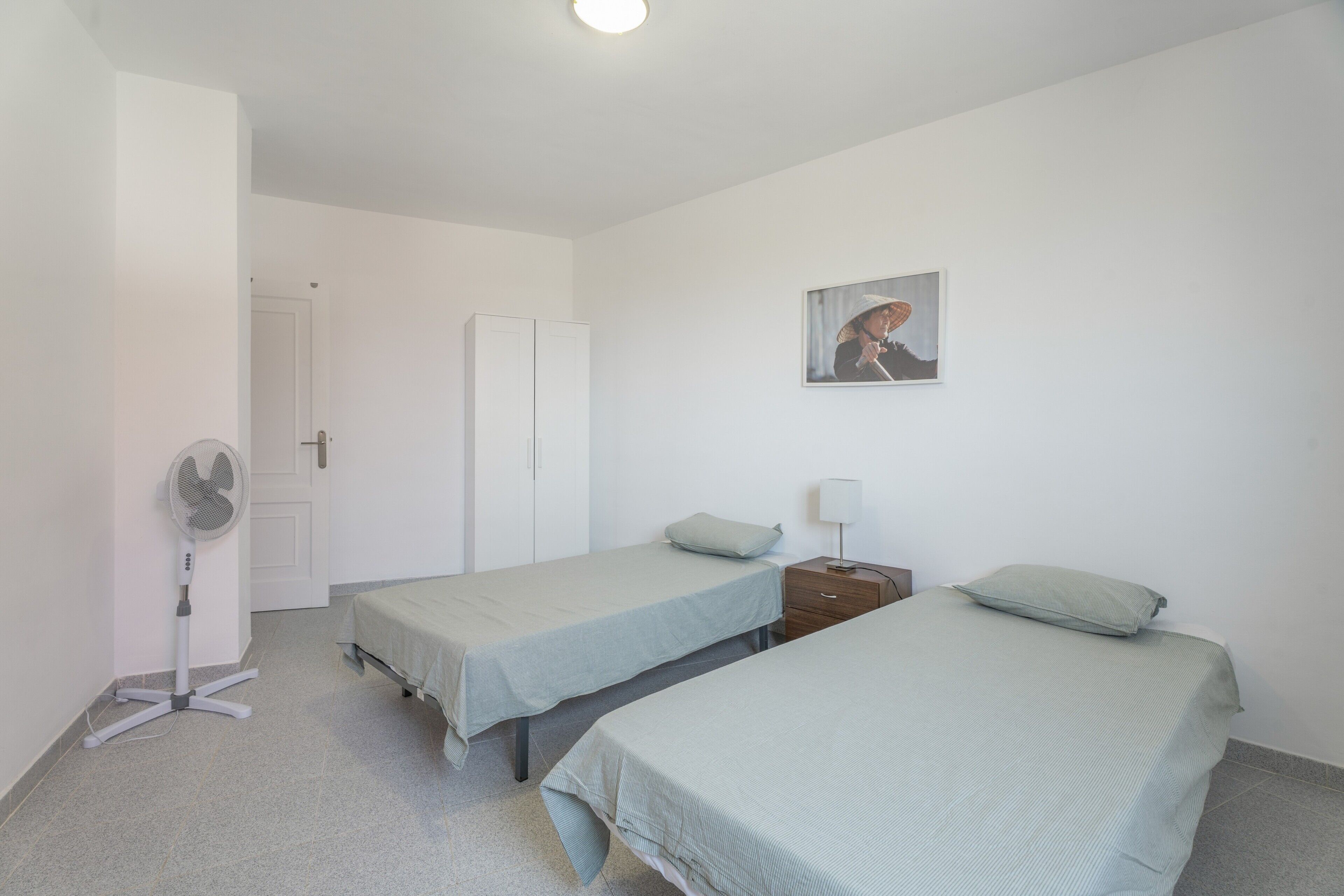 A room with two beds, a standing fan, a door, and a wall-mounted picture.