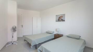 A room with two beds, a standing fan, a door, and a wall-mounted picture.