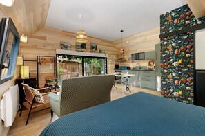 A room with a bed, a chair, a table, a window, and a kitchen area.