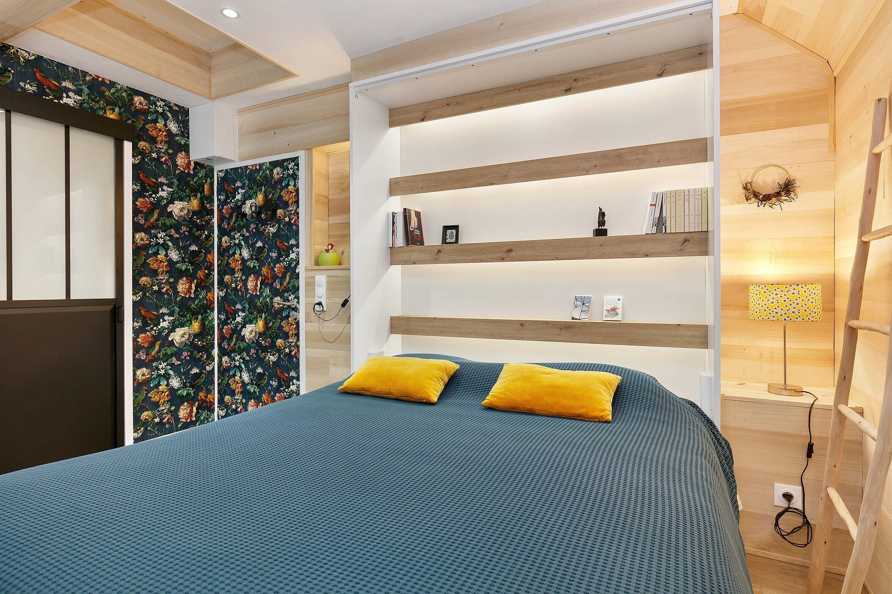 A bedroom with a bed, two yellow pillows, a wooden headboard, and a shelf with books and a lamp.
