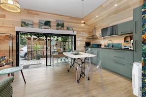 A modern kitchen with wooden flooring, a dining area with a table and chairs, and a view through a glass door to an outdoor patio.