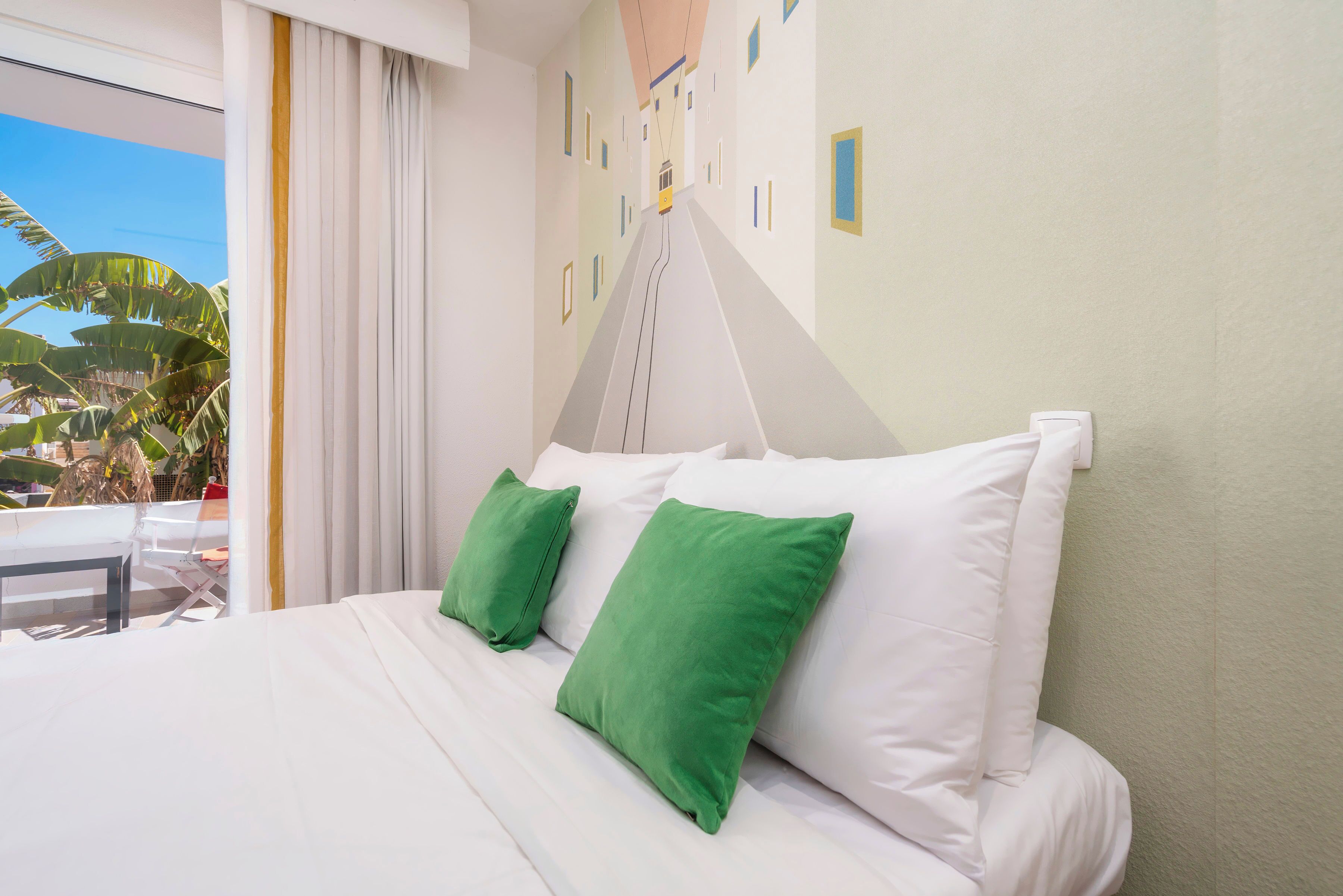 Habitación de hotel con cama, almohadas verdes, ventana con cortinas y vistas a palmeras.