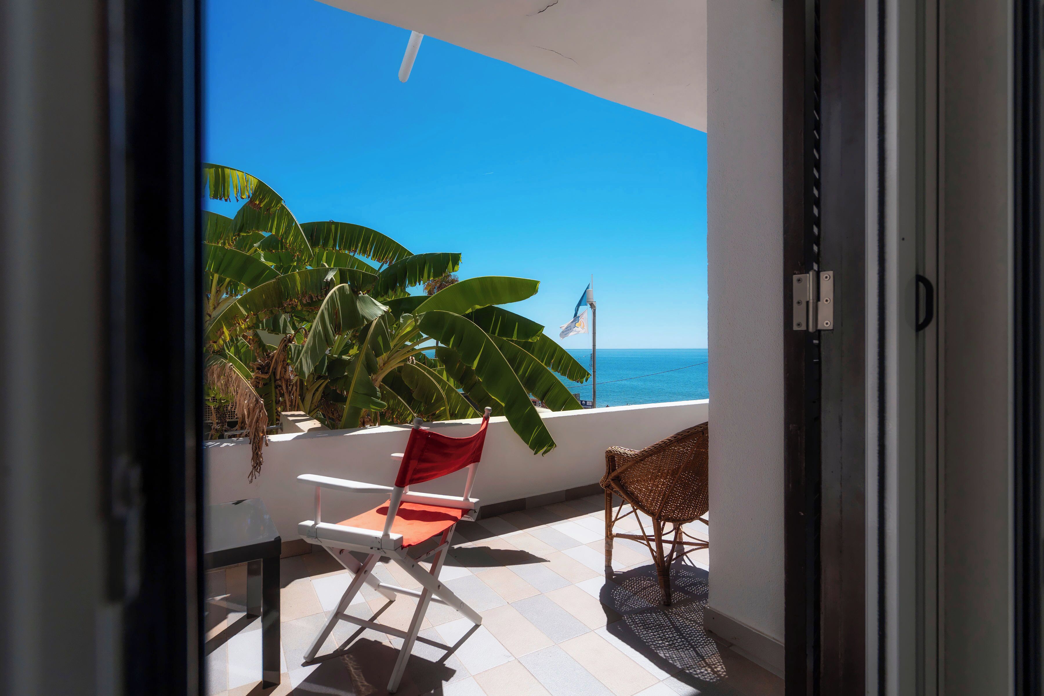 A balcony with a view of the ocean, a red chair, a wicker chair, and a flag.