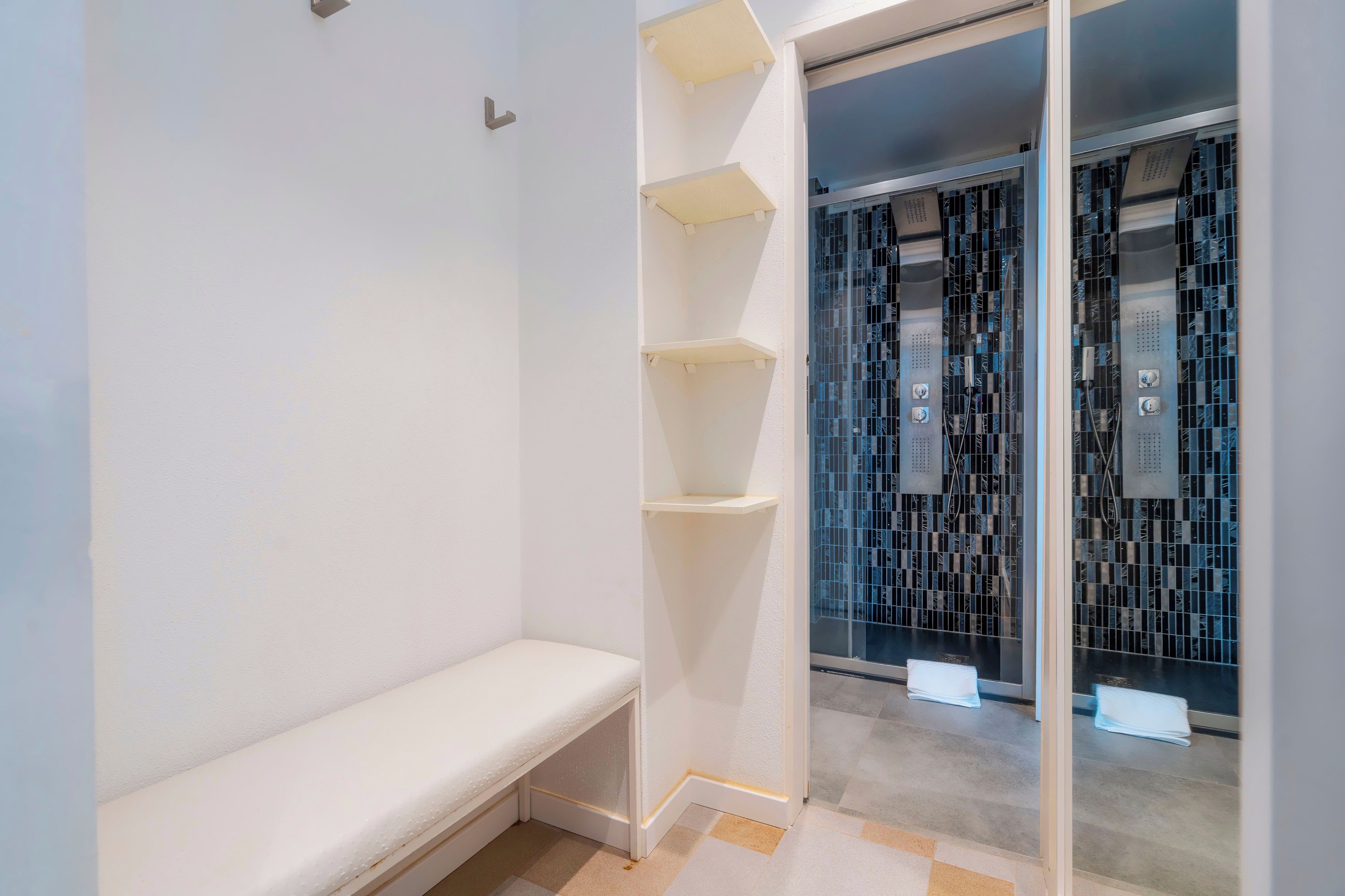 A room with a bench, white shelving, and a glass door leading to a tiled area.
