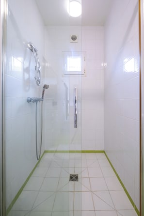 A small, tiled shower room with a glass door, a handheld showerhead, a wall-mounted faucet, and a window above the door.