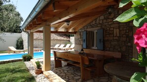 Ein überdachter Außenbereich mit Pool, Liegestühlen, einem Holztisch und Steinwänden.