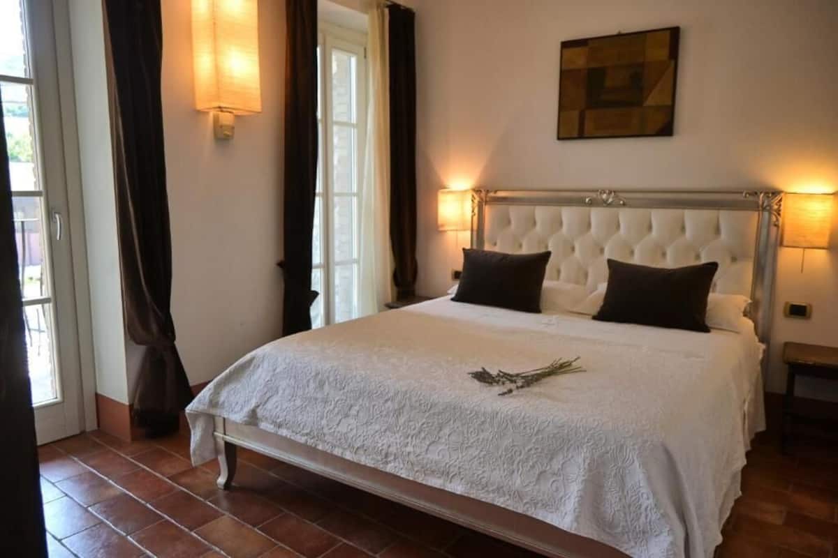 Une chambre à coucher avec un grand lit, deux lampes de chevet, une fenêtre avec des rideaux et une œuvre d’art accrochée au mur.