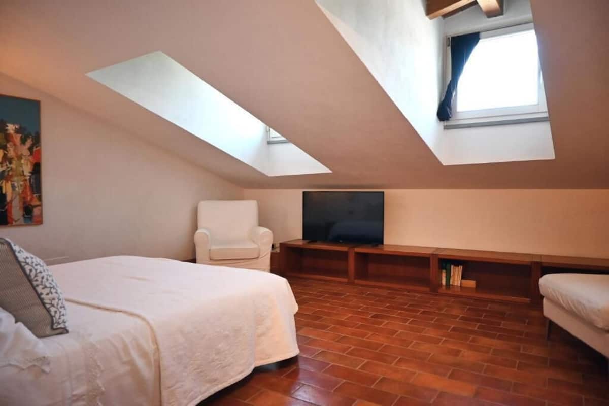 Une chambre avec un plafond en pente, un lit, une chaise, un téléviseur et une fenêtre.