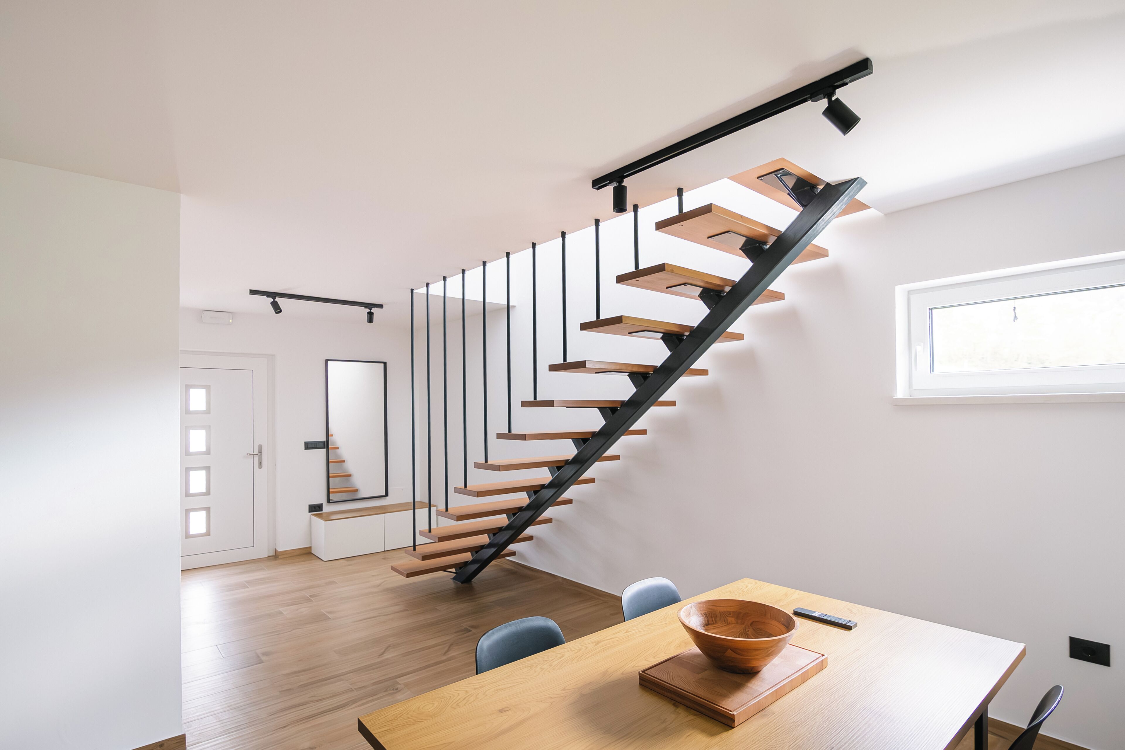 A modern interior with a wooden staircase, a dining area with a wooden table, and a window providing natural light.