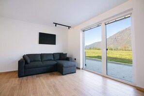 A modern living room with a sectional sofa, a mounted artwork, and a large sliding glass door leading to a scenic outdoor view. - Soča River Resort - Boutique Apartments (Tolmin)