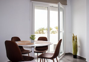A dining area with a round table, six brown chairs, a small potted plant, and a view through a glass door to an outdoor area with palm trees. - Gaviotas Beach Golf (El Ejido)