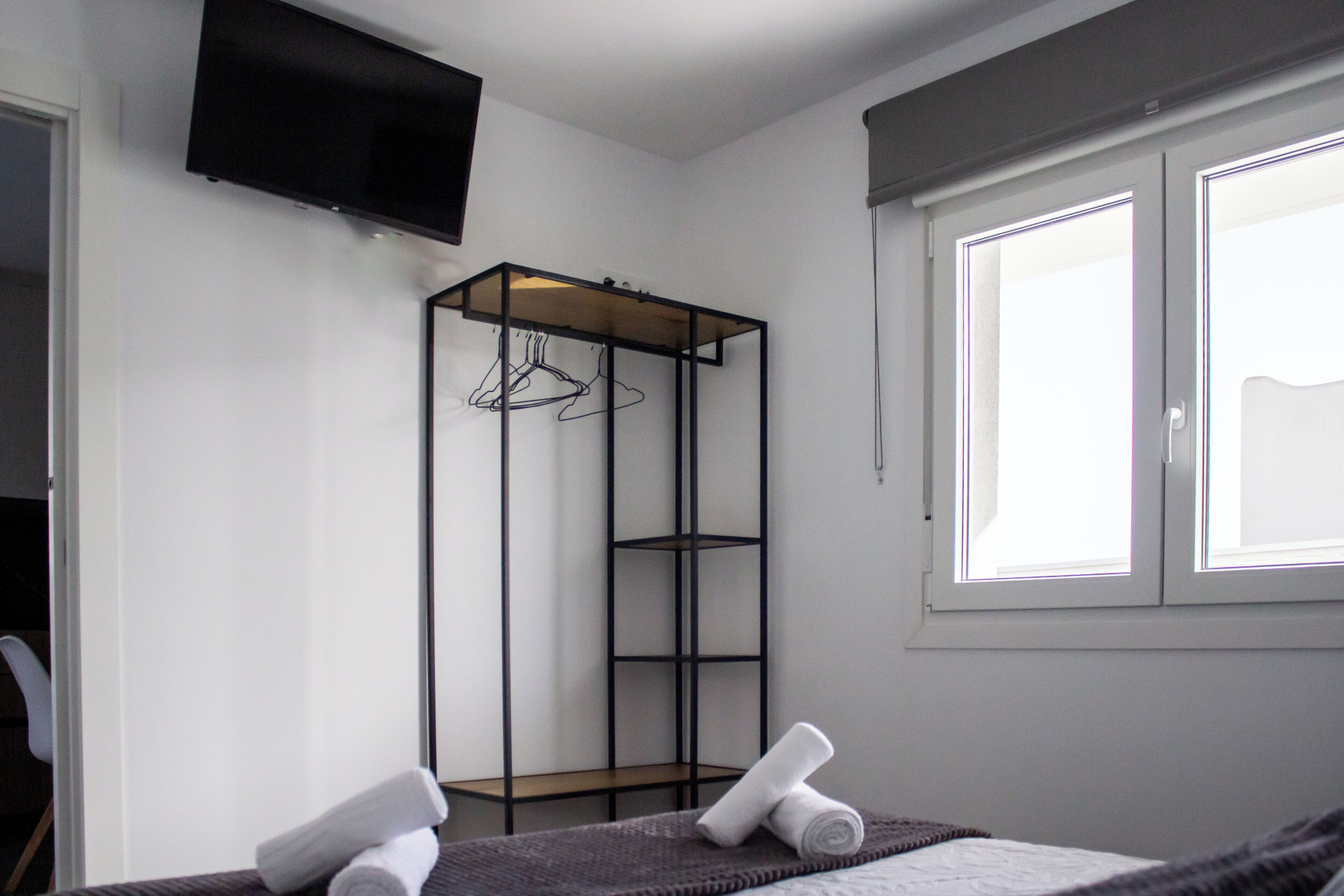 A hotel room with a bed, a TV mounted on the wall, a window with blinds, and a metal wardrobe with shelves.