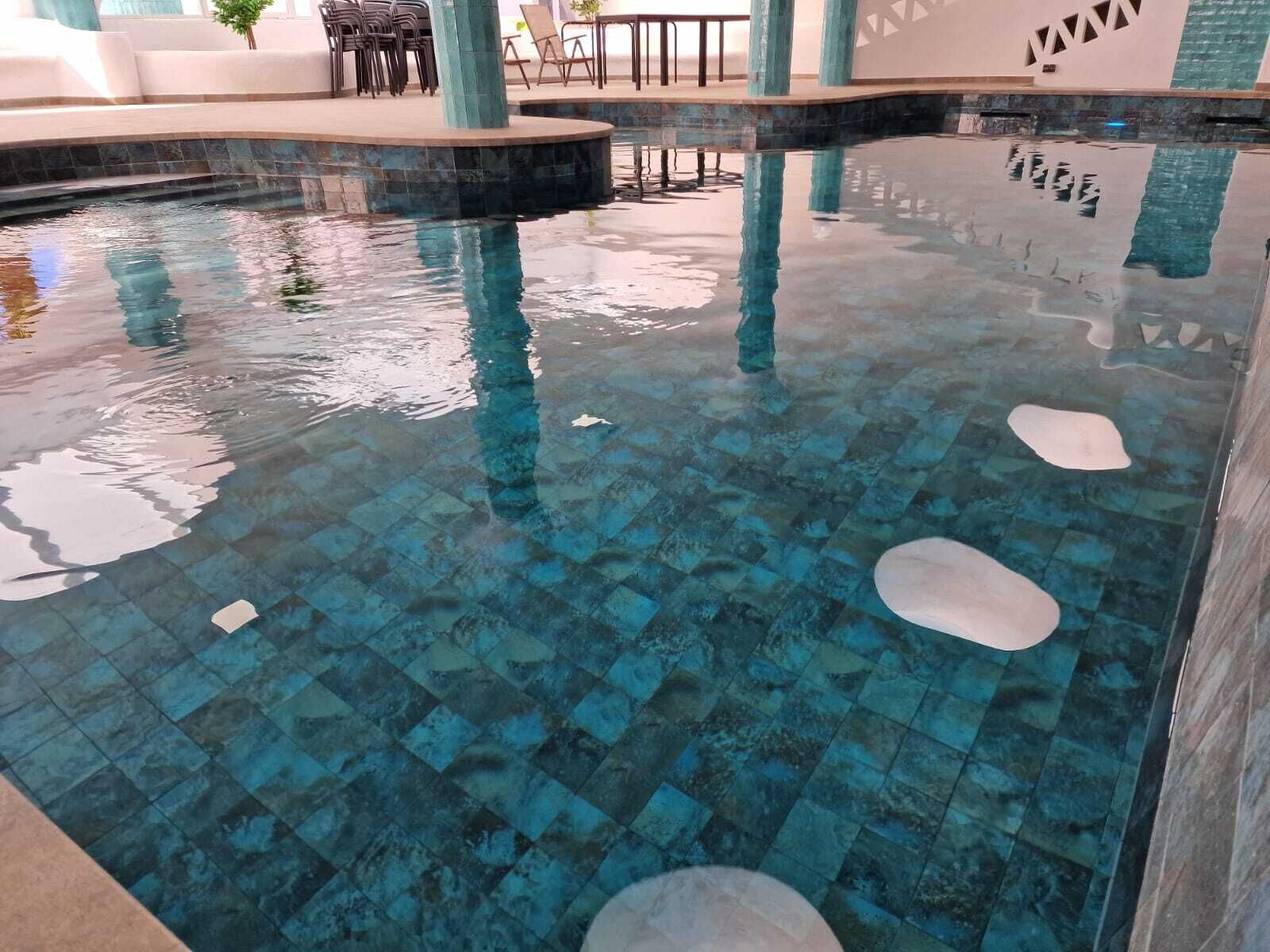 Una piscina interior despejada con suelo de baldosas, sillones de descanso y un área de estar con mesas.