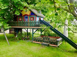 Ein buntes Holzhaus mit Balkon und Rutsche, umgeben von üppigem Grün.