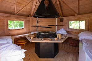 Ein rustikales Blockhaus-Interieur mit zentralem Holzofen, zwei Fenstern und einem Bett mit Kissen.