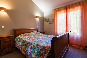 A bedroom with a wooden bed, two bedside tables, a window with sheer curtains, and wall-mounted lights.