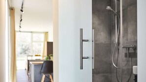 Un baño moderno con ducha, puerta de vidrio y una cocina visible al fondo.