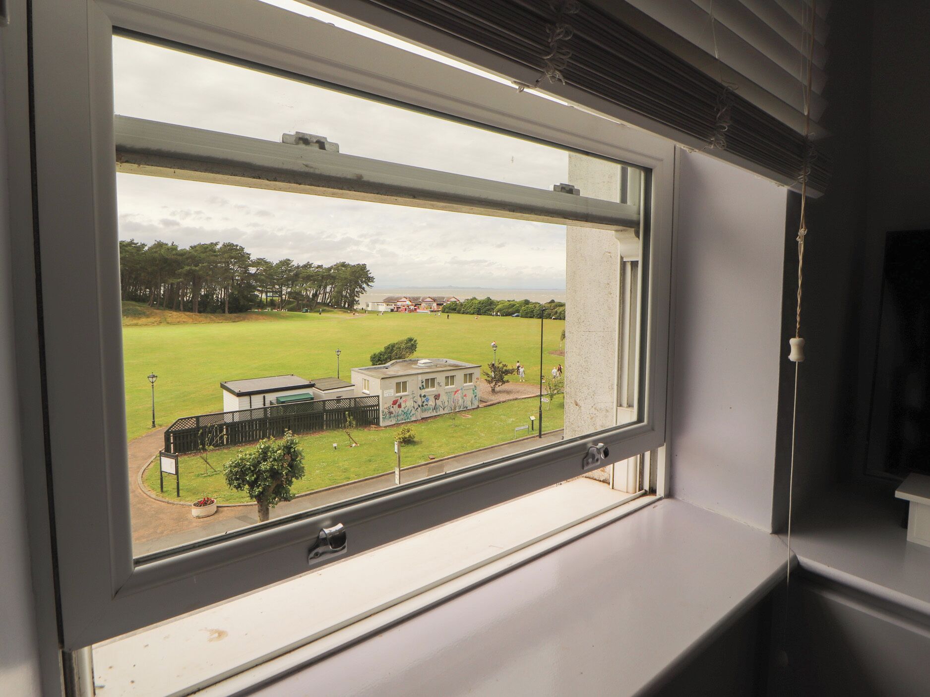 A window with a view of a grassy field, a building with a green roof, and a fence.