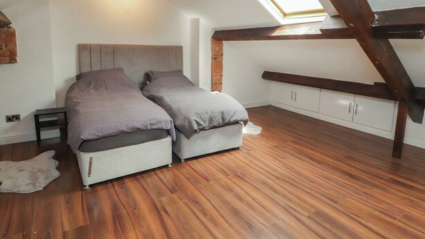A loft bedroom with a sloped ceiling, a skylight, a bed with grey bedding, wooden flooring, and a white wardrobe. - Sugar Loft (Wigton)