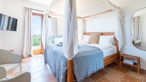 Ein Schlafzimmer mit einem Himmelbett, einem Wand-Flachbildfernseher, einem kleinen Nachttisch und Blick ins Freie durch ein Fenster.