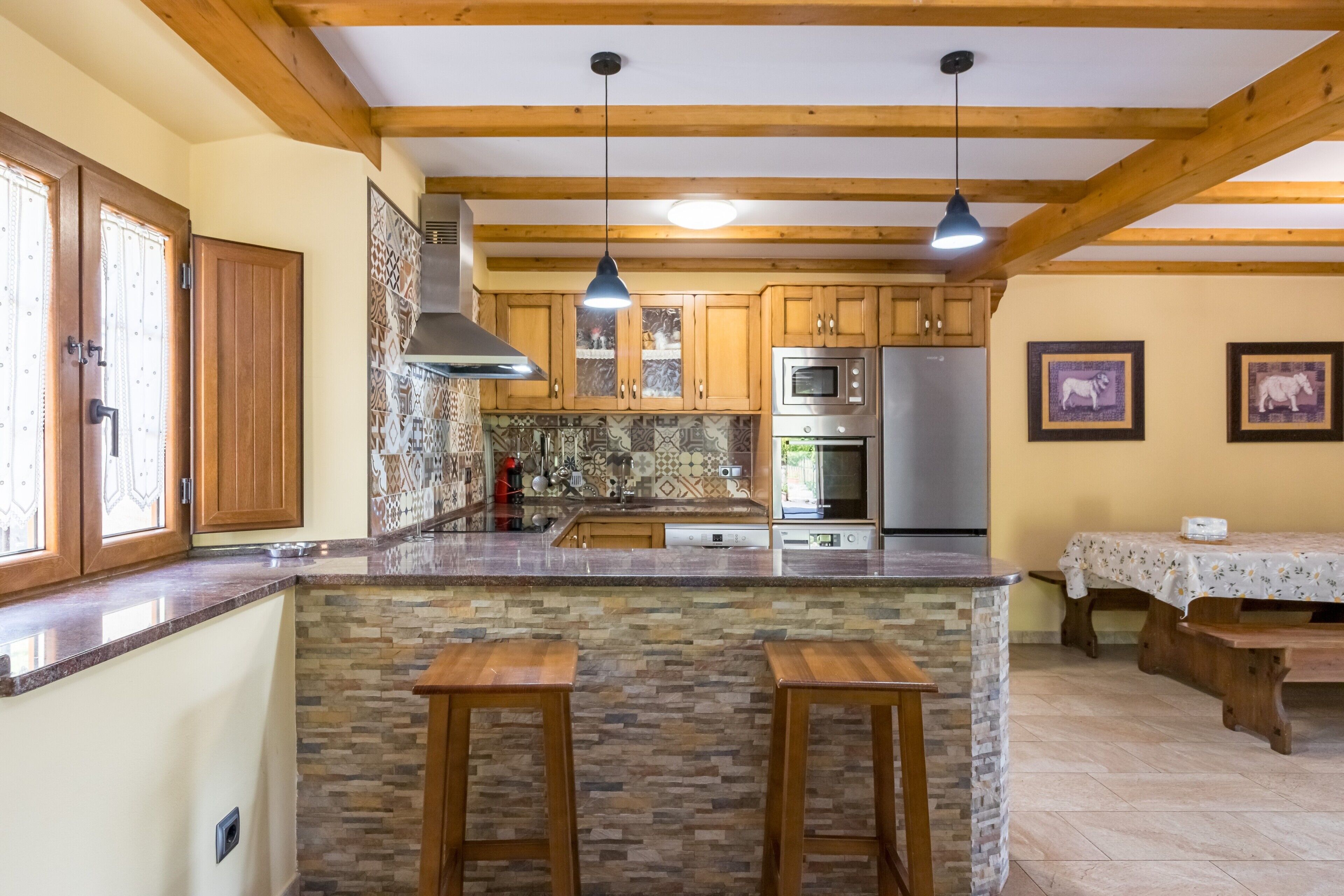 A kitchen with wooden beams, a stone island, two barstools, a dining table with a tablecloth, and a refrigerator.
