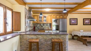 A kitchen with wooden beams, a stone island, two barstools, a dining table with a tablecloth, and a refrigerator.