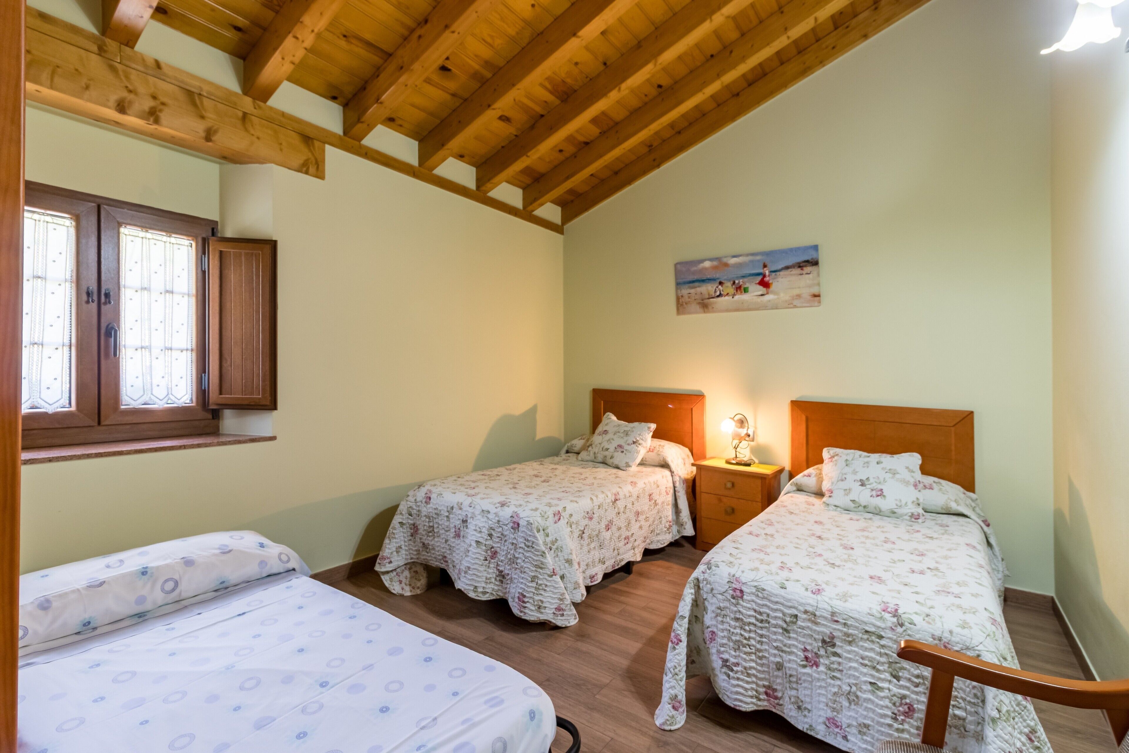 A bedroom with two beds, wooden beams, a window with blinds, a painting on the wall, and a nightstand with a lamp.