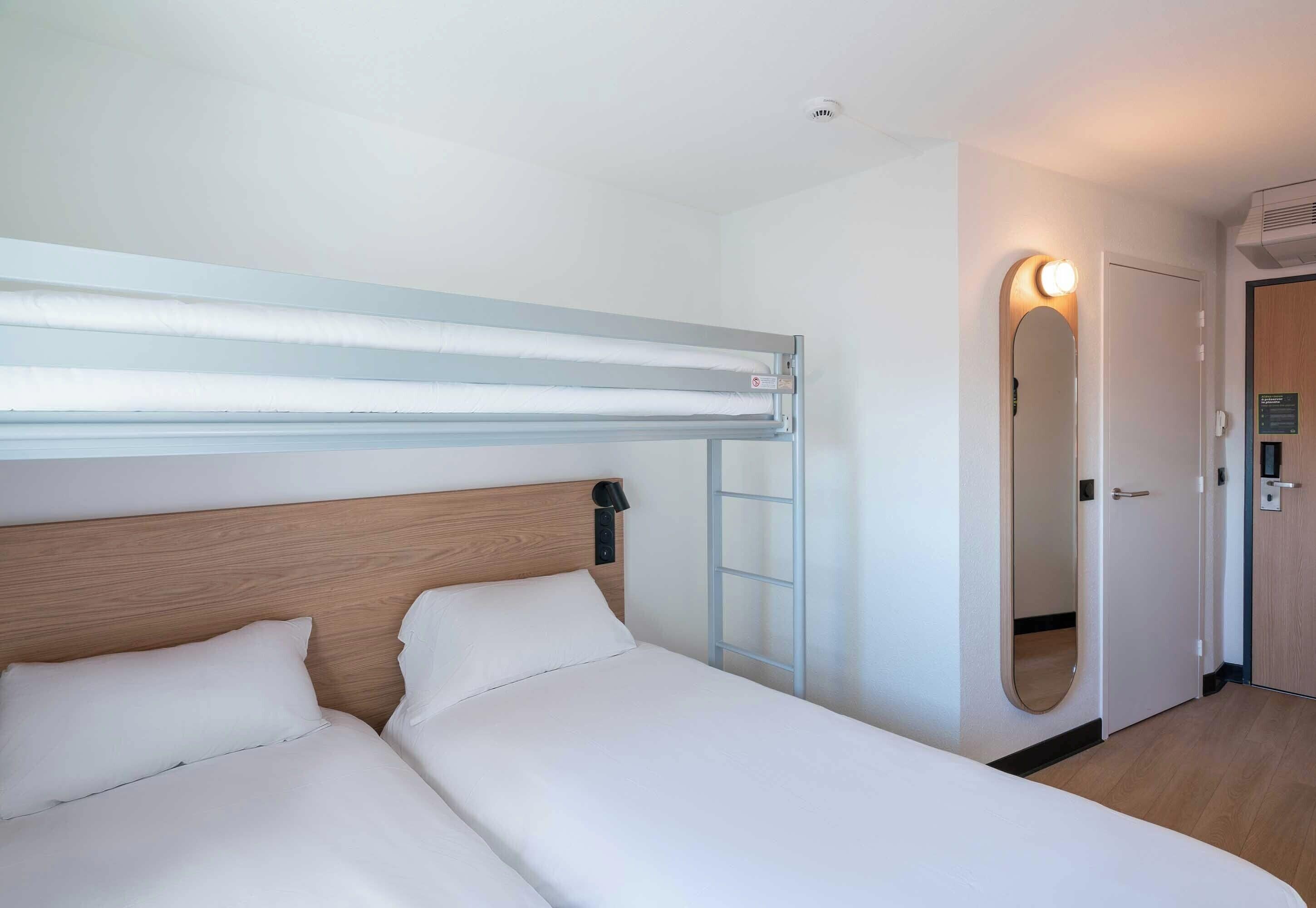 A hotel room with a bunk bed, a bedside table with a lamp, a mirror, and a door with a light switch.