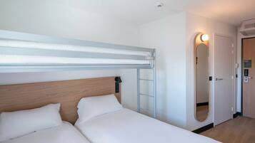 Habitación de hotel con una cama litera, una mesita de noche con lámpara, un espejo y una puerta con interruptor de luz.