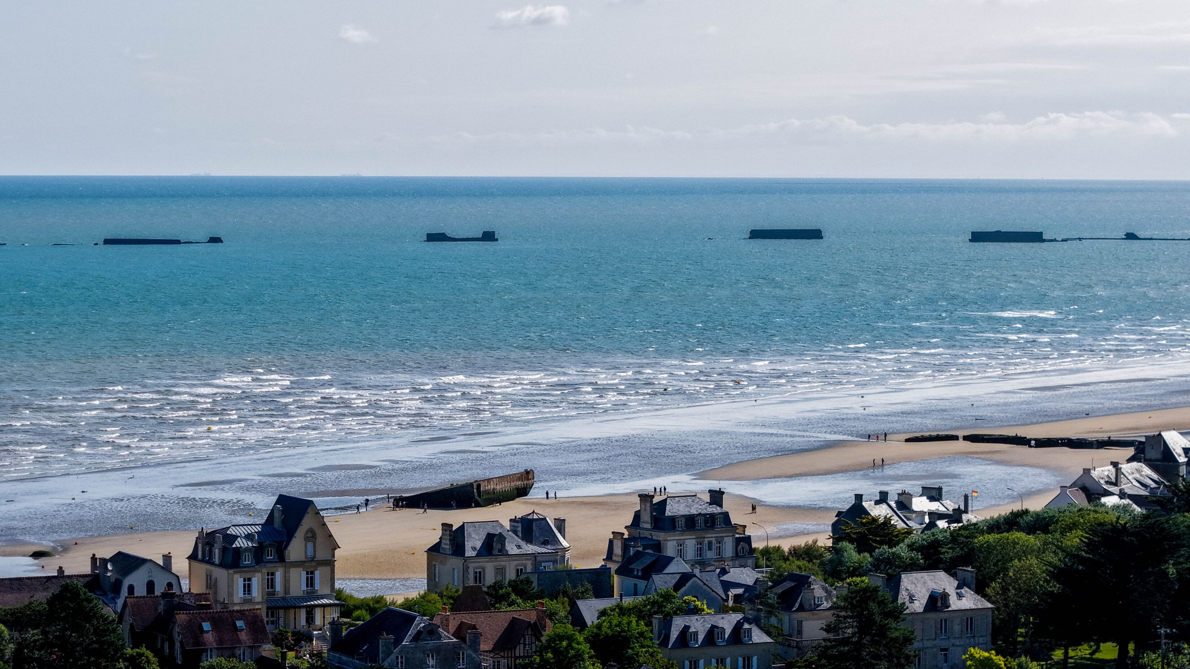 A coastal town with a beach, several ships in the distance, and historic buildings.
