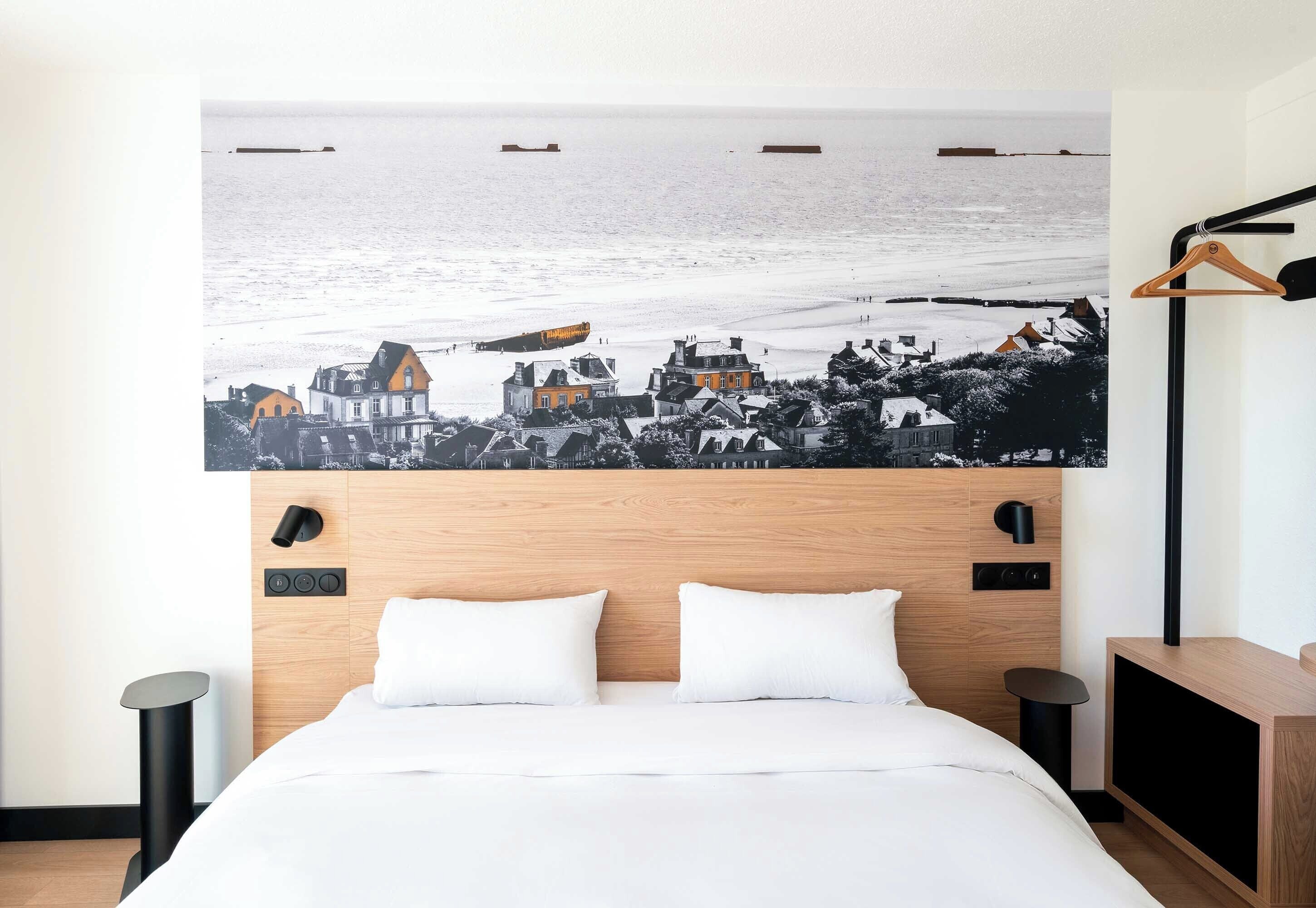 A hotel room with a bed, a headboard with controls, a wall-mounted artwork of a coastal scene, and a wooden nightstand.