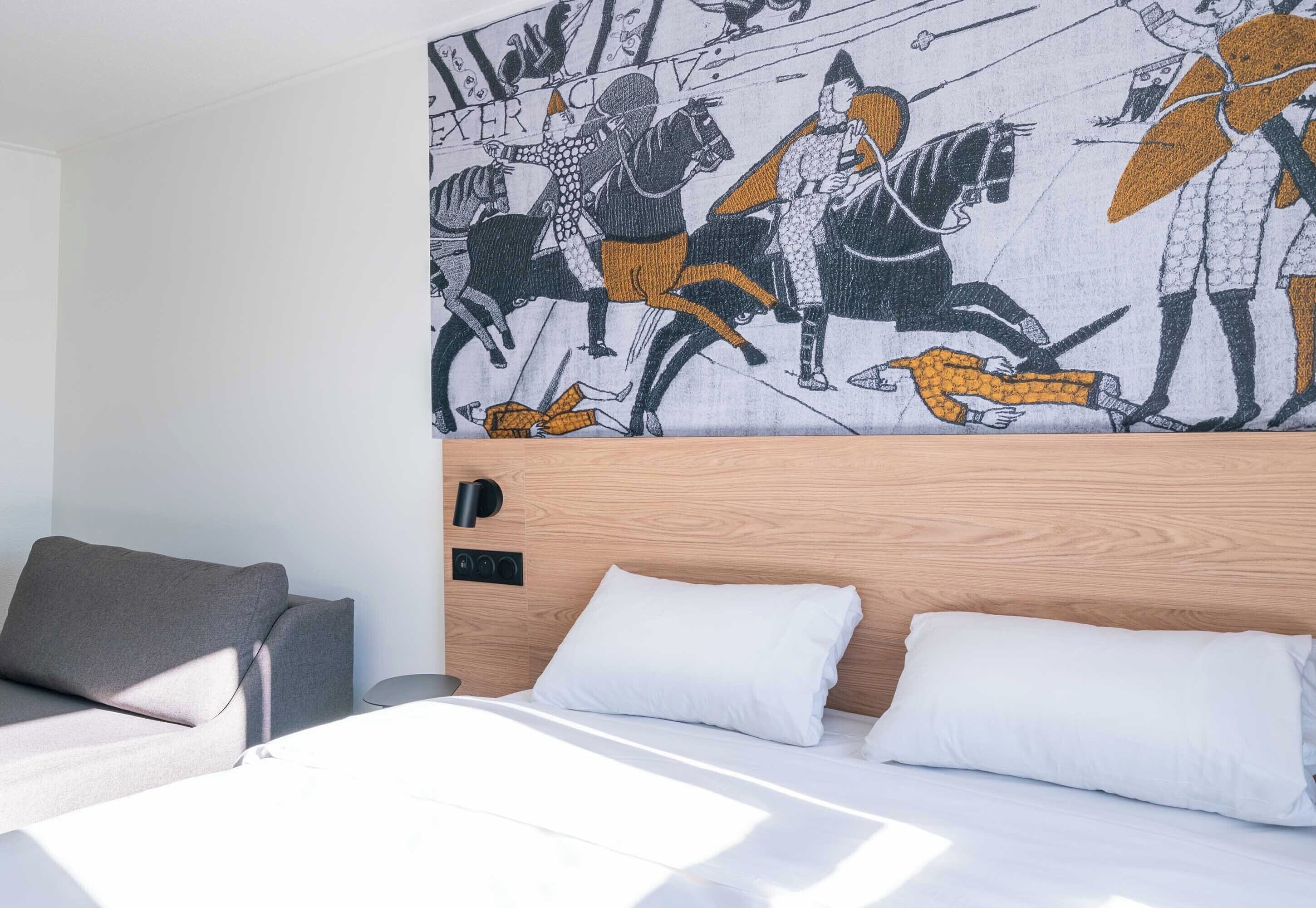 A modern hotel room with a wooden headboard, a bed with white linens, a grey sofa, and a wall mural depicting a medieval battle scene.