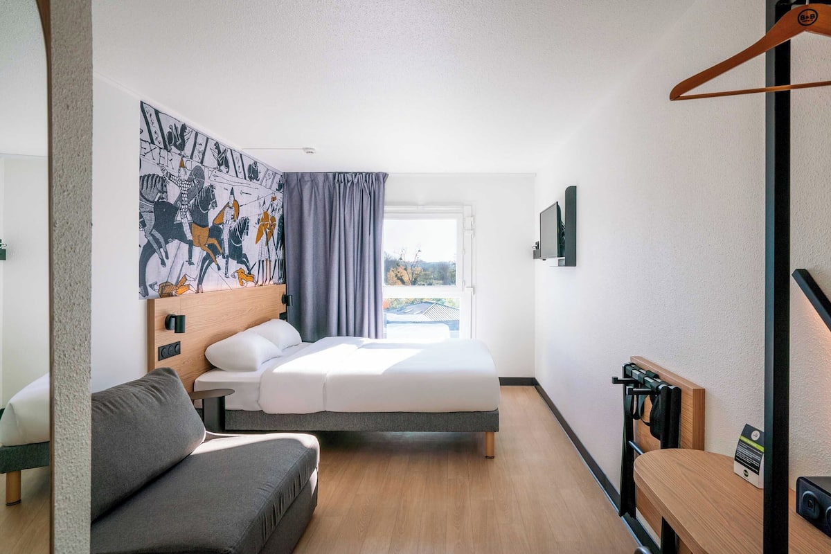 Une chambre d’hôtel avec un lit, un canapé, une tête de lit en bois, une fenêtre avec des rideaux et une vue sur l’extérieur.