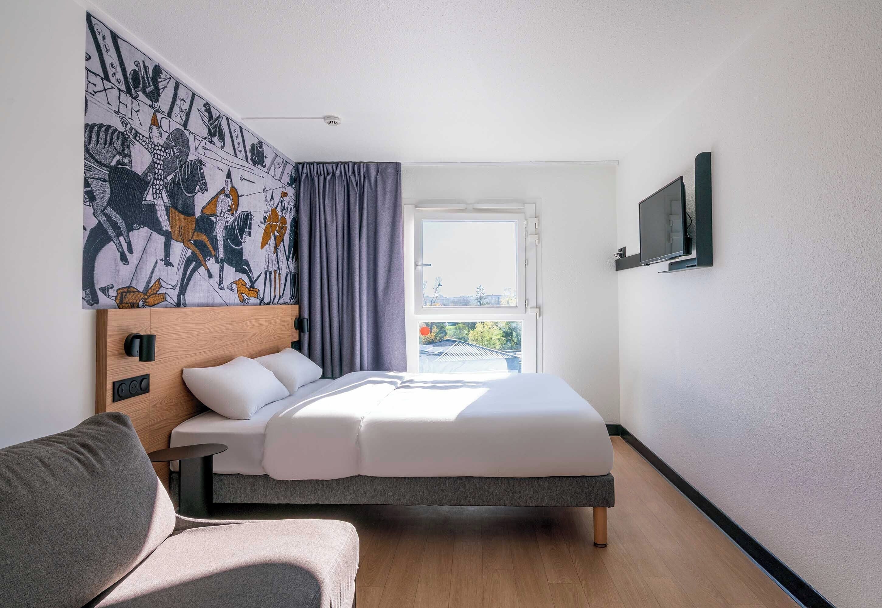 A modern hotel room with a bed, a sofa, a large window, and a wall-mounted TV.