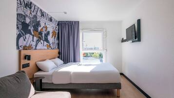 A modern hotel room with a bed, a sofa, a large window, and a wall-mounted TV.