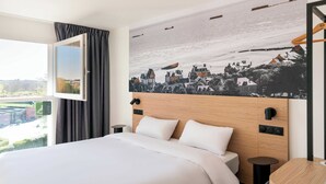 Ett hotellrum med en säng, en sänggavel med en väggmålning som föreställer en stadsbild, ett fönster med gardiner och utsikt över omgivningarna.