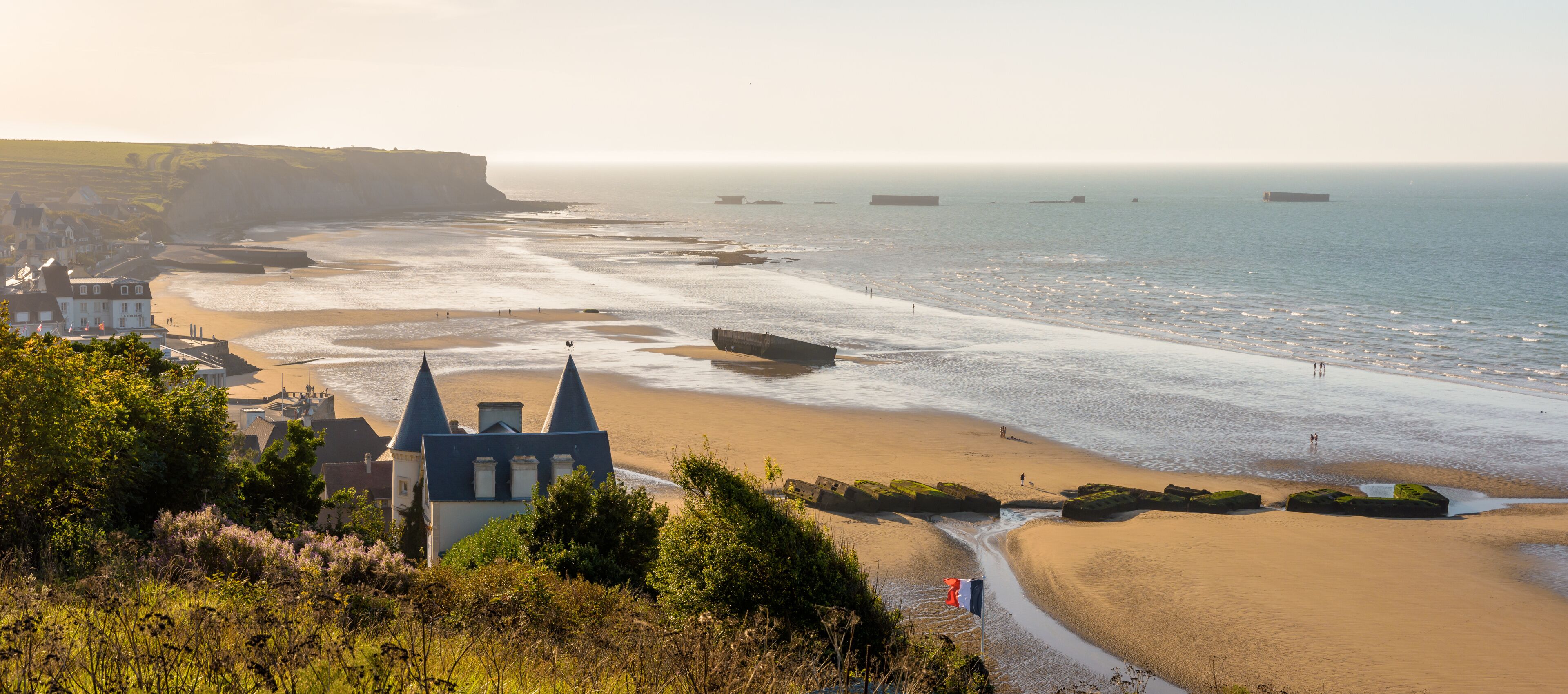 A coastal town with a prominent building featuring a pointed roof, a sandy beach, and a calm sea.