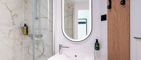 A modern bathroom with a glass shower enclosure, a white sink, a mirror, and a wooden door.