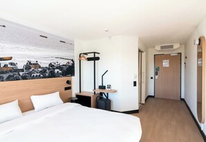 Quarto de hotel com cama, escrivaninha, cadeira, cabideiro e porta.