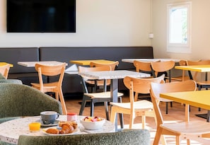 Un espace repas avec des tables et des chaises, un tĂ©lĂ©viseur fixĂ© au mur et une fenĂȘtre laissant entrer la lumiĂšre naturelle.