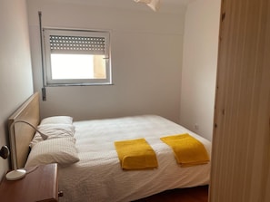 Ein kleines, ordentlich bezogenes Bett mit weißer Bettdecke und zwei gelben Handtüchern, einer Nachttischlampe und einem Fenster mit Jalousien.