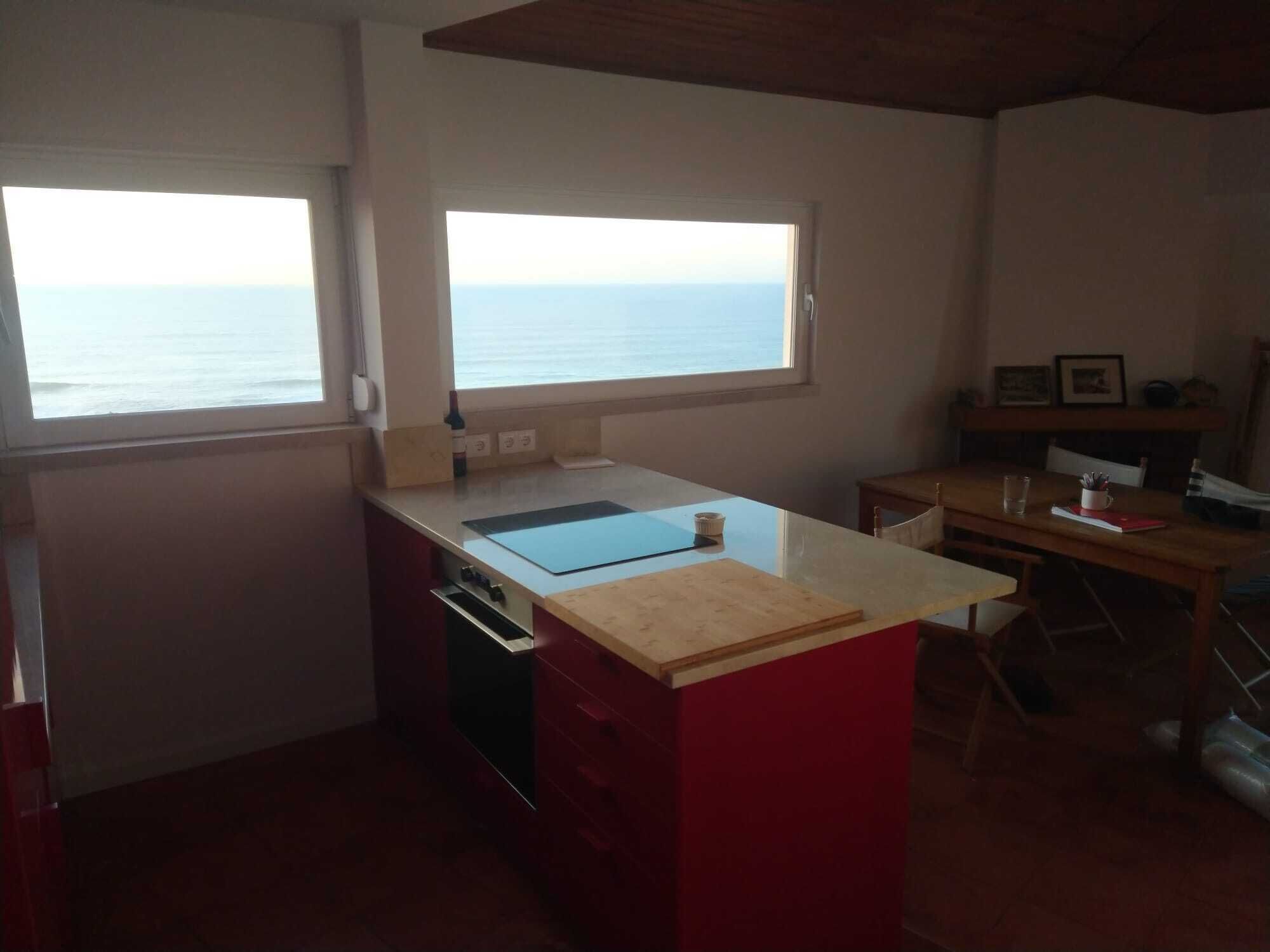 A kitchen with a view of the sea, a wooden table, and a red cabinet.